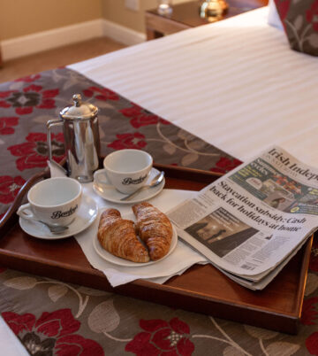 Hotel room with breakfast tray, croissants, coffee, and newspaper on a floral bedspread.