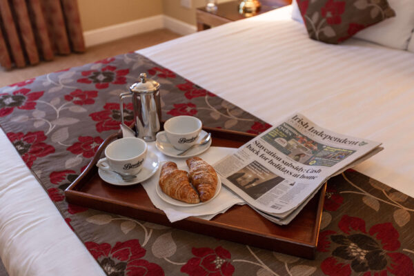 Hotel room with breakfast tray, croissants, coffee, and newspaper on a floral bedspread.