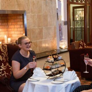 Two women enjoy afternoon tea by a cosy fireplace at Hamlet Court Hotel.