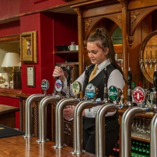 Woman serving drinks at Hamlet Court Hotel bar with warm wooden decor and cosy atmosphere.