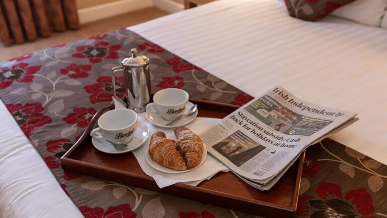 Breakfast tray with croissants, coffee, and newspaper on a cosy hotel bedspread.