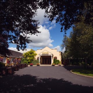 Entrance of Hamlet Court Hotel surrounded by lush greenery under a bright blue sky.
