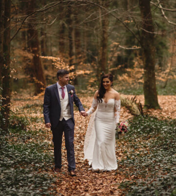 Couple walking hand in hand through a wooded path in autumn, surrounded by fallen leaves.