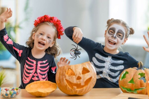 Two children in Halloween costumes carving pumpkins in a cosy, bright room.