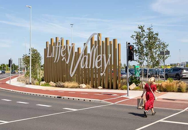 Woman crossing street near Liffey Valley sign on sunny day.